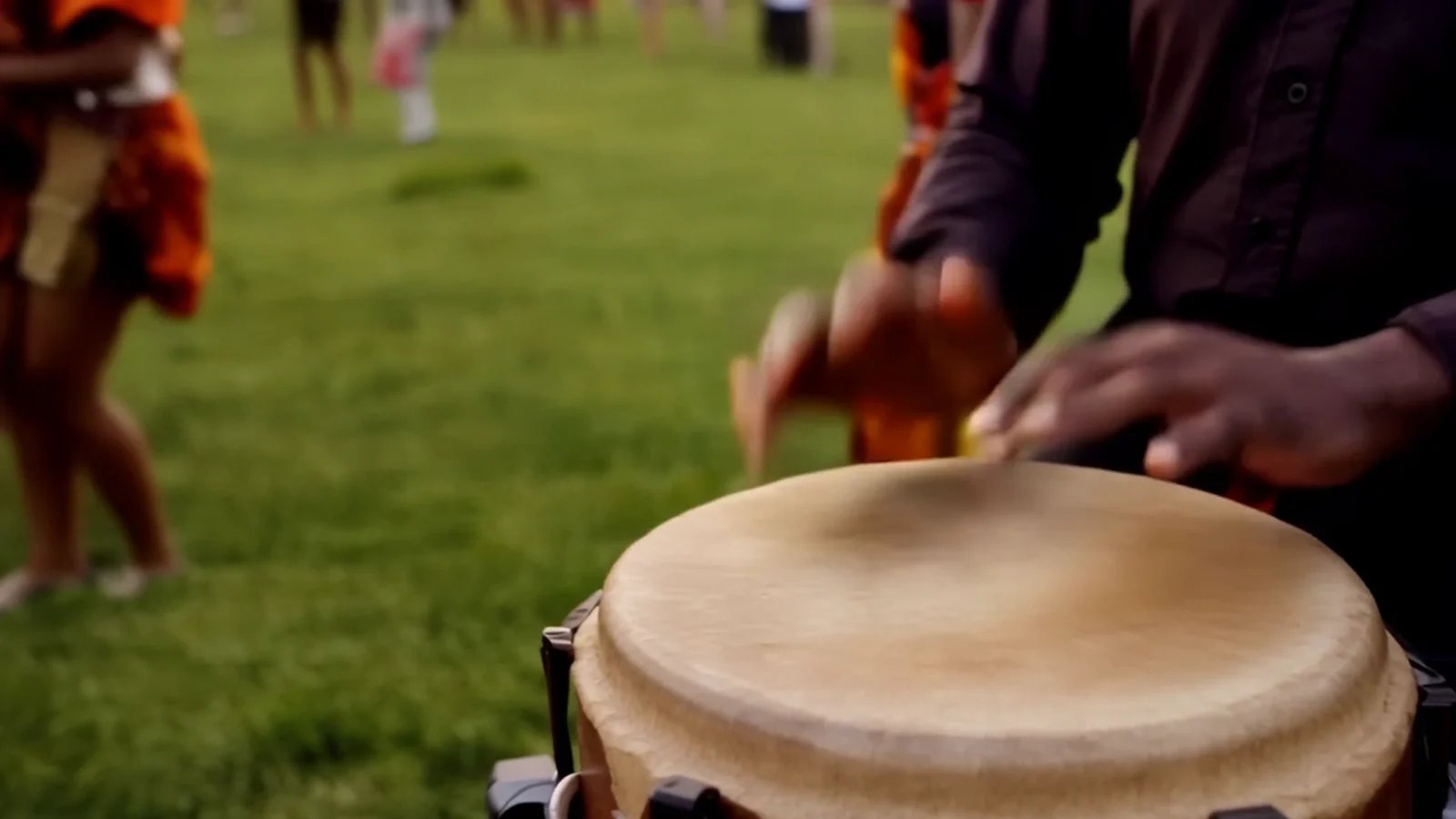 Hands playing a hand drum on a grassy festival field, with dancers blurred in the background