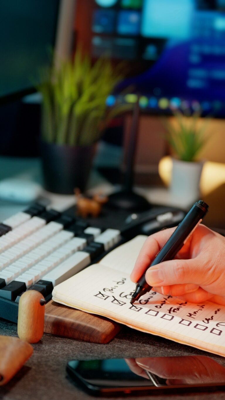 A hand writing a checklist with checkboxes in a notebook next to a keyboard